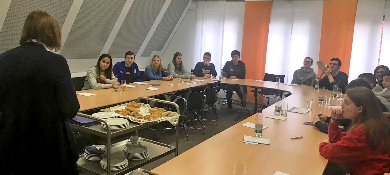 Schüler sitzen im Rechteck an Tischen, vor ihnen eine Sprecherin, ein Tablett mit Gebäck