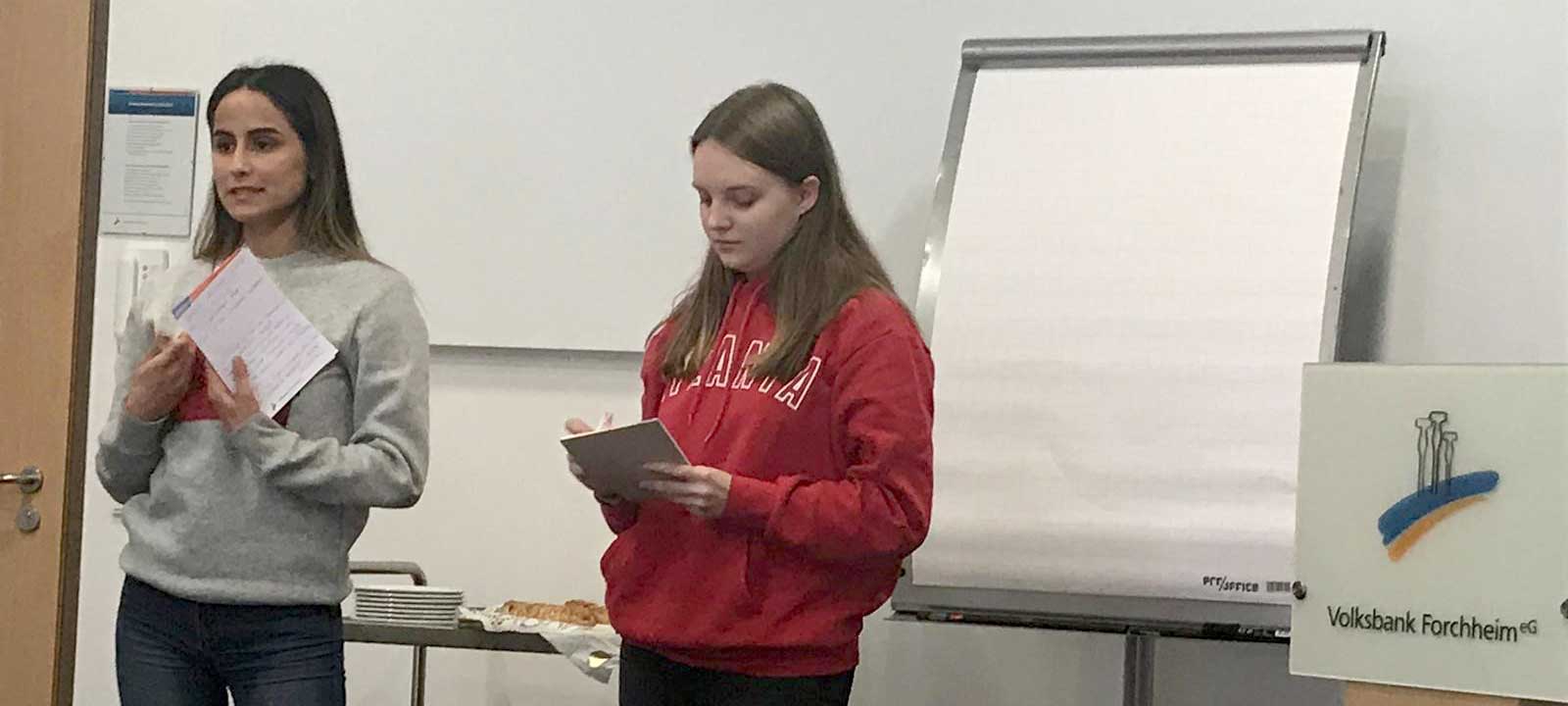 Schülerinnen stehen mit Schreibblöcken vor einer Tafel und sprechen