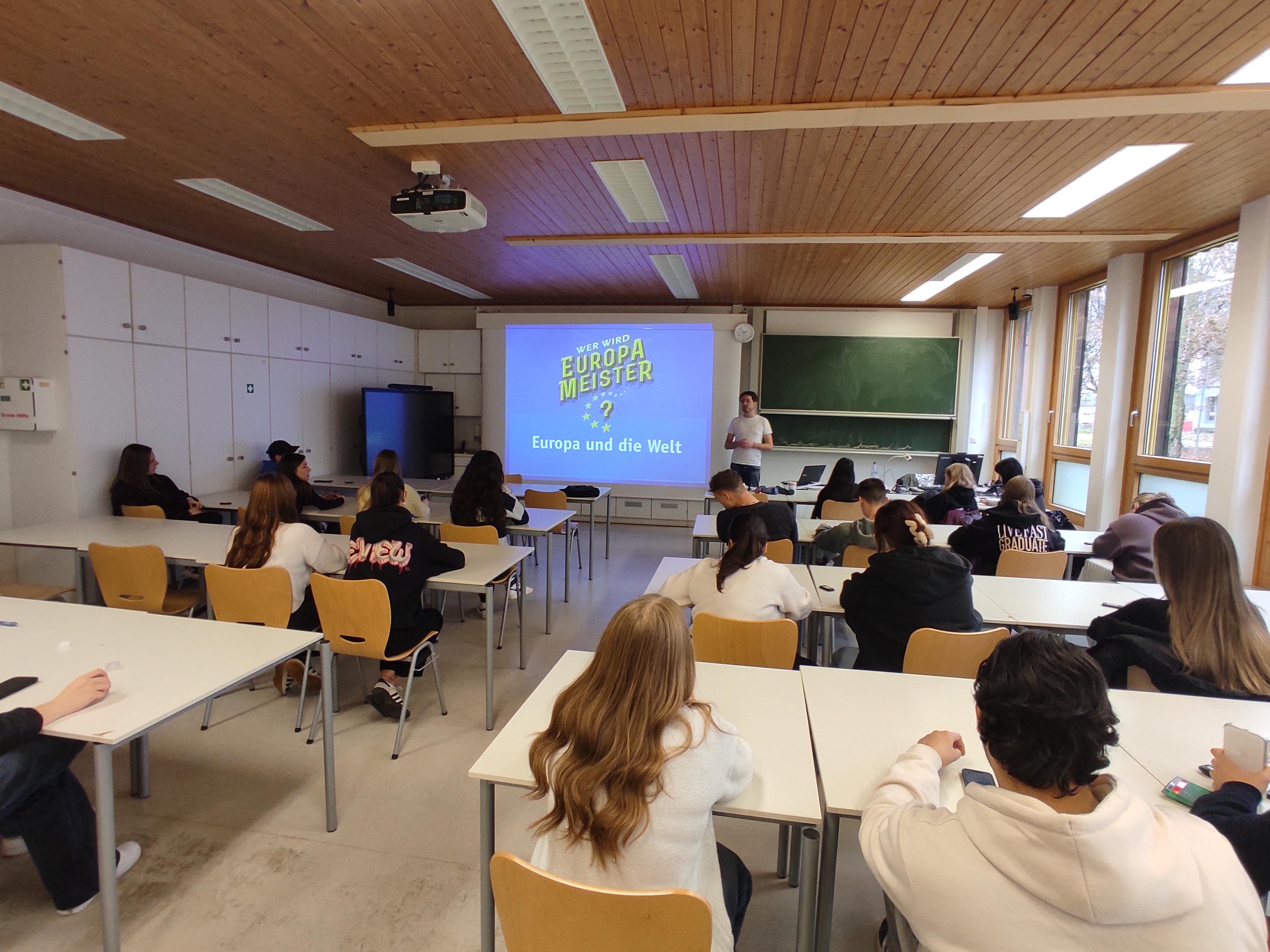 Vortragender neben Touchpanel, sitzende Schülerinnen und Schüler